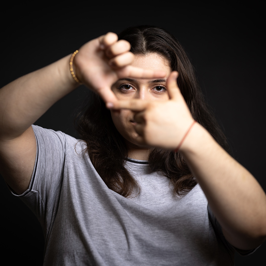 Alessia - Be Extraordinary campaign - she is holding her hands up in front of her face making a square like a frame. She is wearing a grey t-shirt and gold bracelet, standing against a black background.