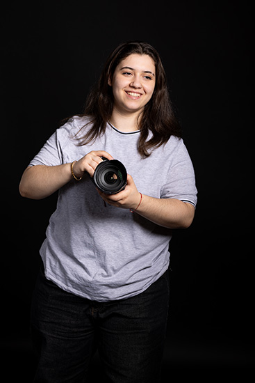 Alessia - Be Extraordinary campaign - she is holding a camera and has long dark hair. She is wearing a grey t-shirt and gold bracelet, standing against a black background.