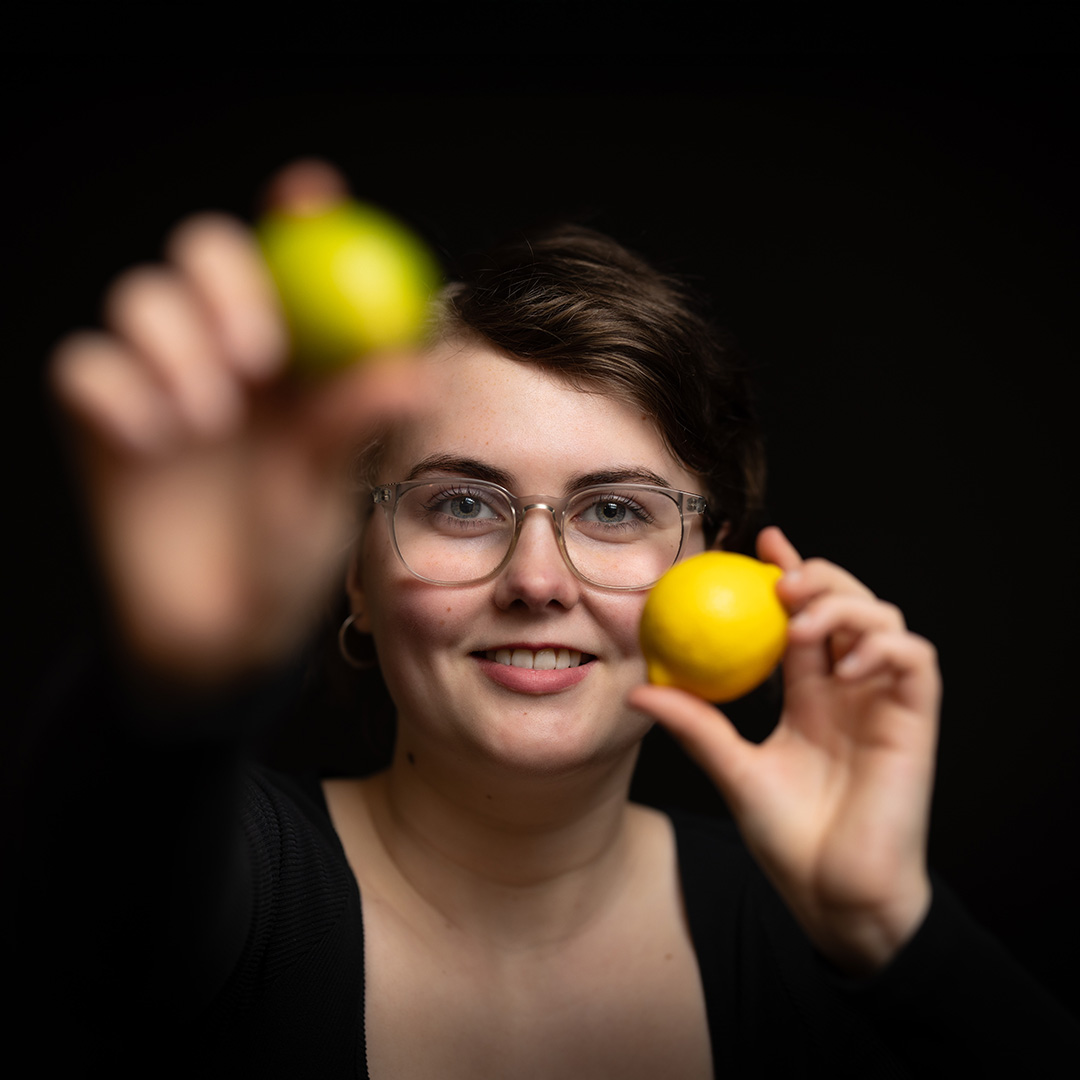Lucy - a Richmond upon Thames professional cookery student - is holding up a lime and lemon in her hands. She is wearing glasses and there is a black background behind her.