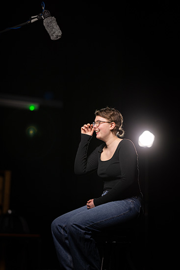 Lucy - a Richmond upon Thames professional cookery student - is sitting on a chair. There is a light on a stand behind her and a mic and boom above her. She is holding her hand up to her glasses.