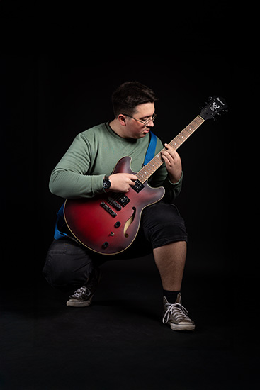 Luka - a Richmond upon Thames Music Performance & Production student - is holding a guitar on his knee and bending down. He is wearing a green long sleeved t-shirt, glasses and sneakers.