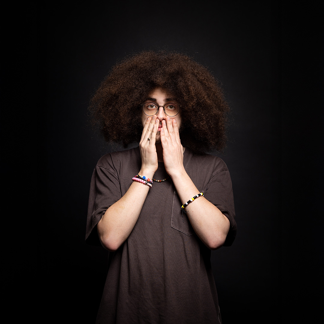Zak - Richmond upon Thames College International A-Level student - he has both hands held up to his face, covering his mouth. He has a a long afro, round glasses and is wearing bead braclets and necklace. 