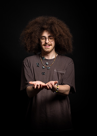 Zak - Richmond upon Thames College International A-Level student - is throwing dice up in the air. He has a a long afro, goatee beard, round glasses and is wearing bead braclets and necklace.
