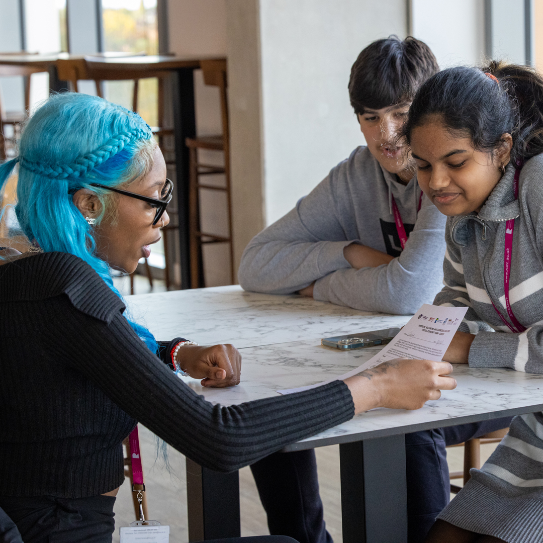 Richmond upon Thames catering students sitting in The View Restaurant talking. One of the students has long blue hair and two others are wearing grey tops and pink lanyards. 