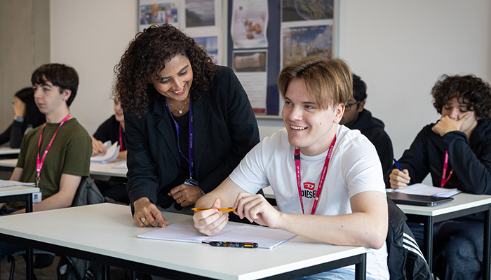 male student sitting at a desk in a classroom, smiling, with female teacher standing by him and smiling too