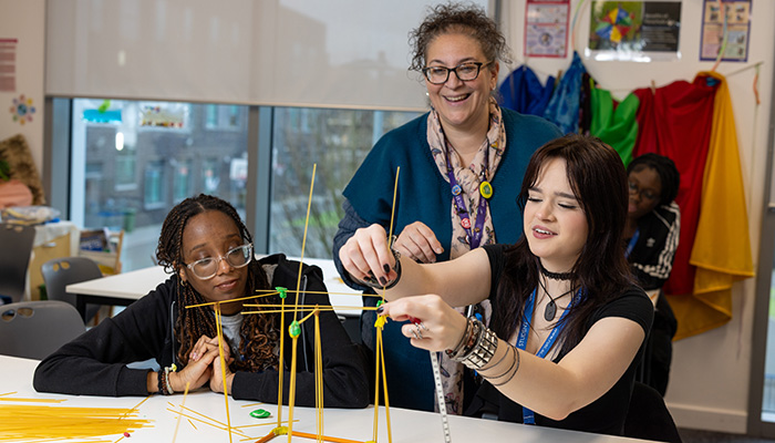 two female students working together to make a structure from marshmallows and spaghetti, with teacher standing behind them smiling with them