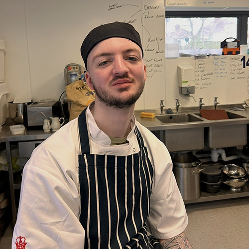 Alfie Green - alumni student from Richmond upon Thames College (RuTC) wearing his chef uniform, stripe apron and hat, sitting in a commercial kitchen setting. There are sinks behind him.