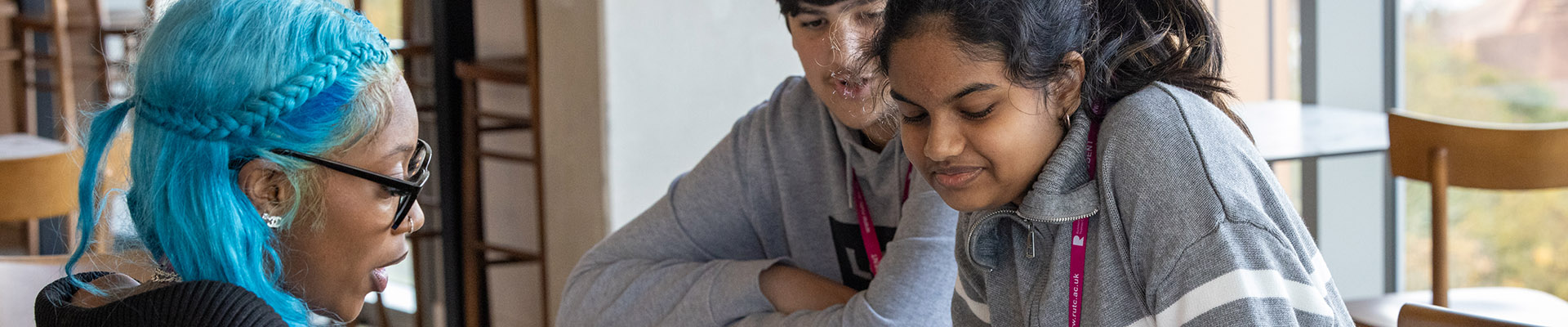 Richmond upon Thames catering students sitting in The View Restaurant talking. One of the students has long blue hair and two others are wearing grey tops and pink lanyards. 