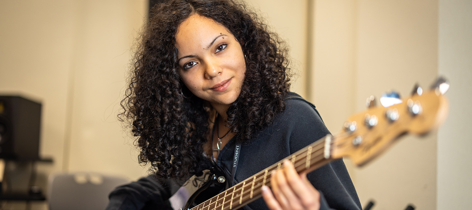 A female Richmond upon Thames College music student in the studio sitting holding a guitar. She has curly sholder length hair and is wearing a black top.