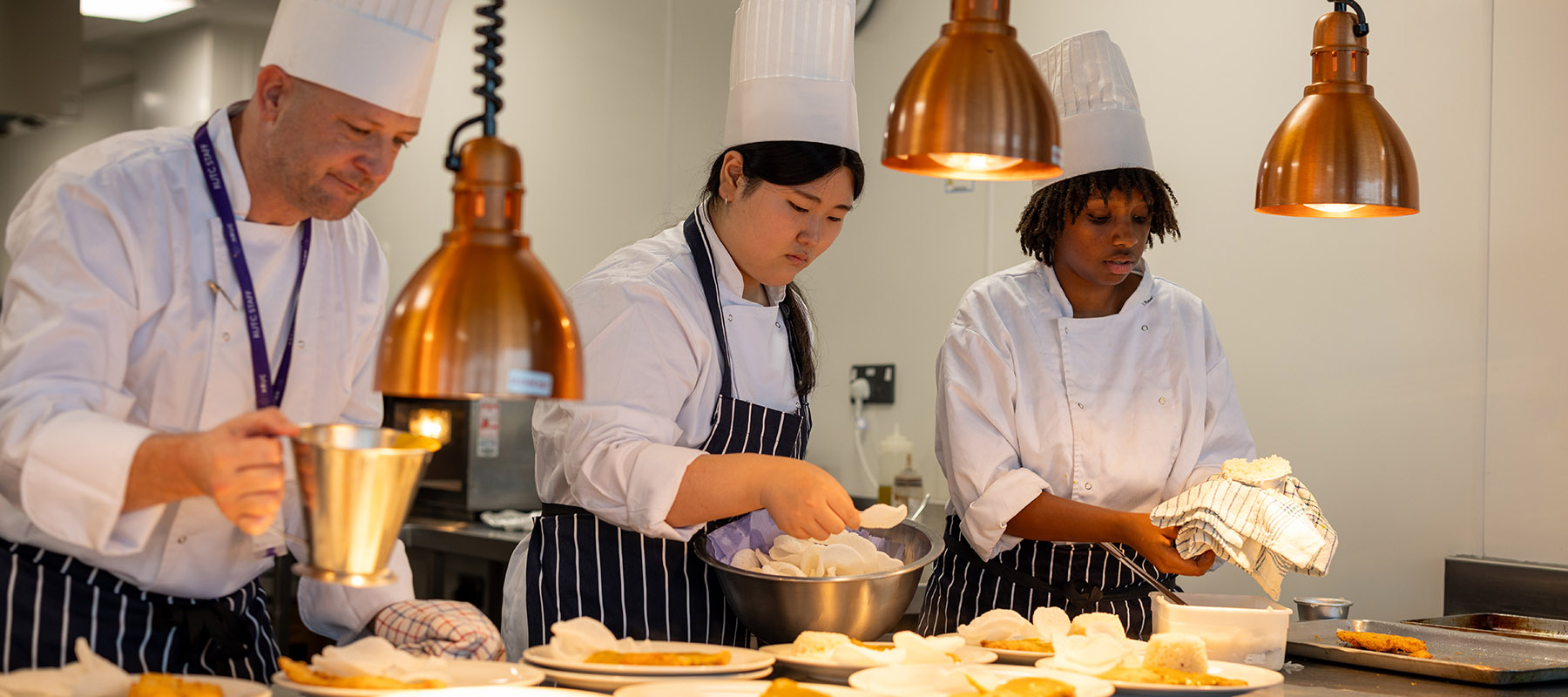 Richmond upon Thames College Professional Cookery staff and students at the pass, plating up food on multiple plates under heat lamps. They are wearing chef hats and uniforms.