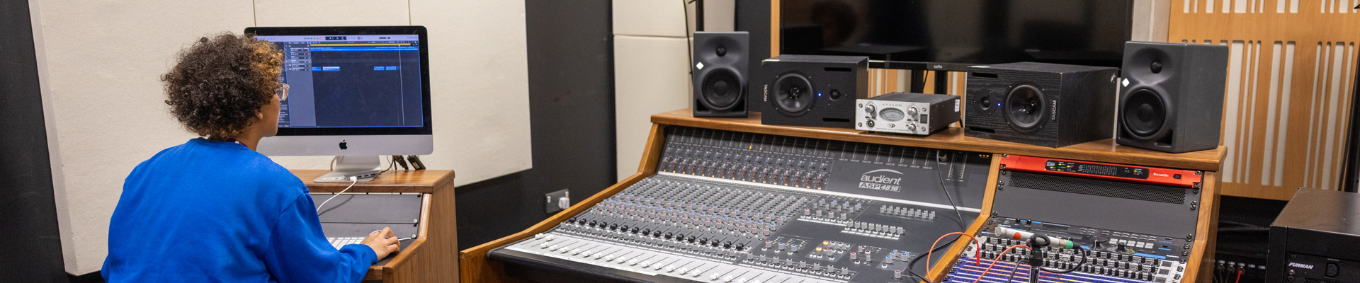 student on mac with back to camera in music studio with large PA system