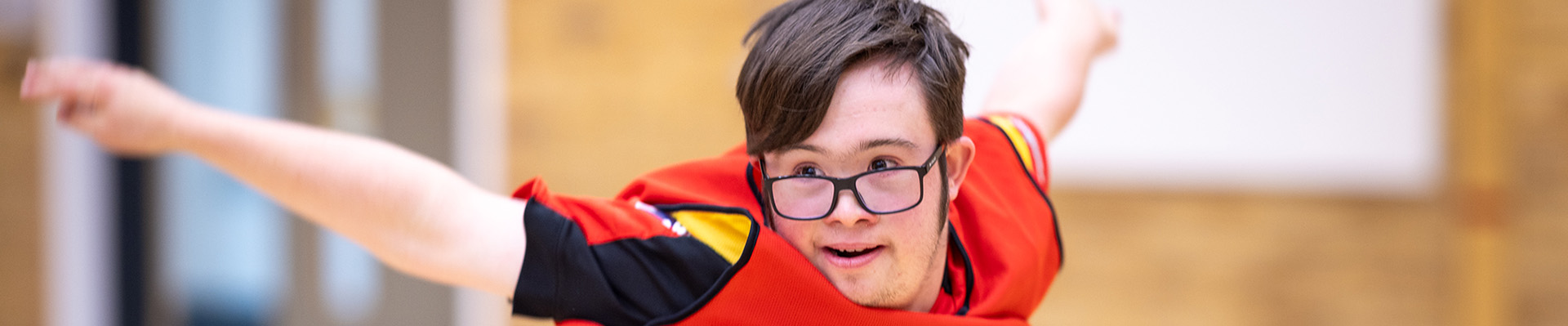 male student with glasses in orange sports vest, jumping, with arms out-stretched, smiling and looking to the left