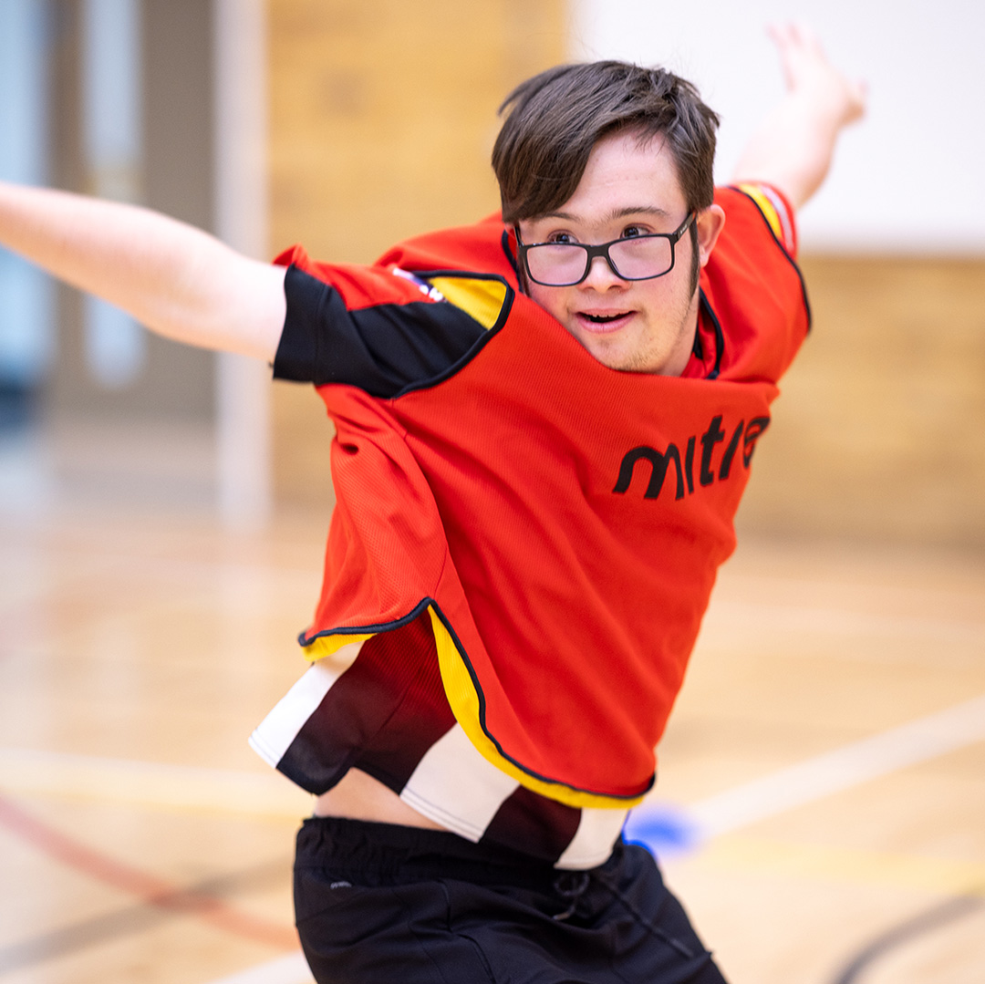male student with glasses in orange sports vest, jumping, with arms out-stretched, smiling and looking to the left