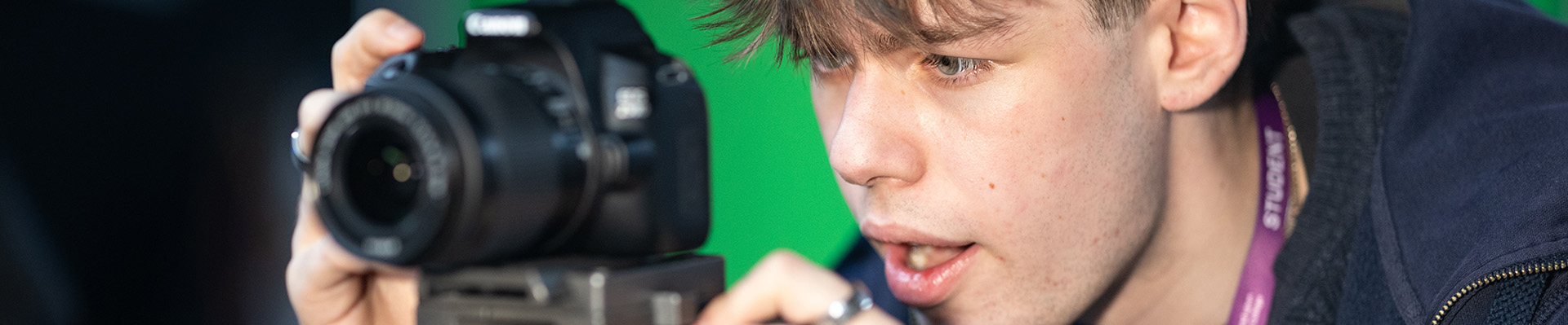 male student looking through the display screen of a DSLR camera with green screen in the background