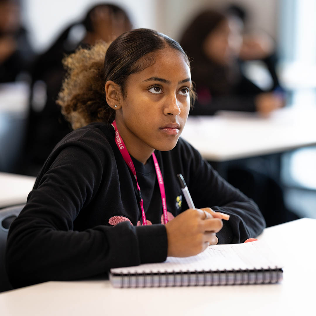 Female Richmond upon Thames student looking up to the front of the class, listening. She is holding a pen over a note pad ready to write. 