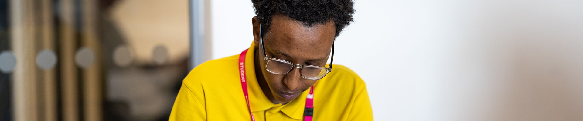 Richmond upon Thames College student studying in a classroom, looking down and writing on a note pad. He is wearing a bright yellow polo shirt, glasses and a pink lanyard.