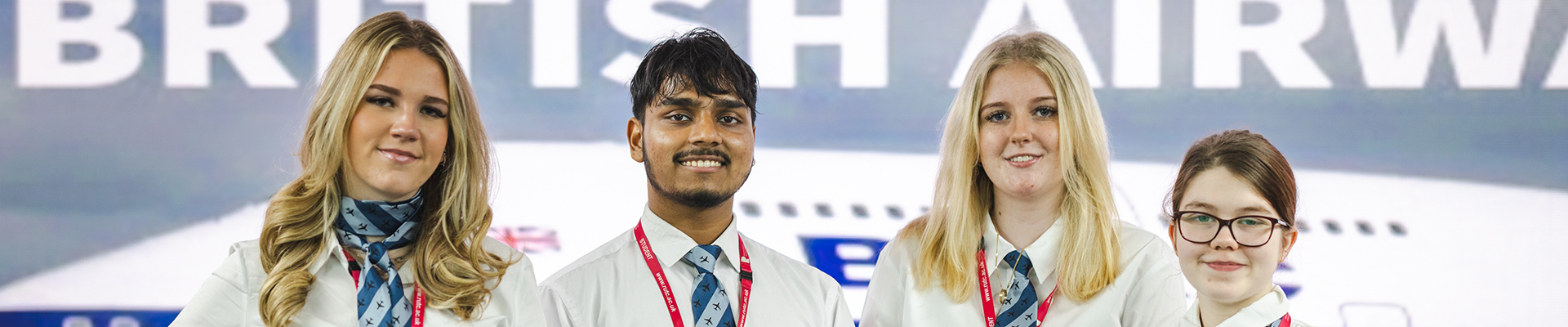 Richmond upon Thames Travel and Tourism students smiling at the camera with a British Airways ad behind them. They are wearing thier new uniforms with blue and white scarfs and ties.