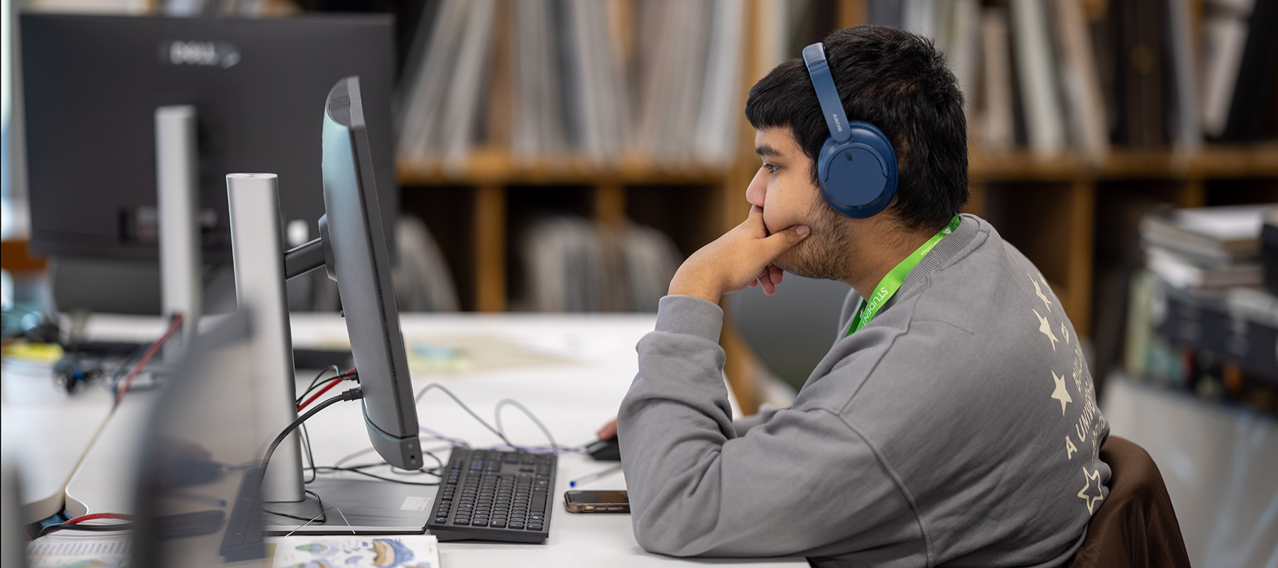 Male Richmond upon Thames student sitting at a computer with blue headphones on. His hand his held up to his mouth and he is wearing a grey top with a lime green lanyard around his neck. 