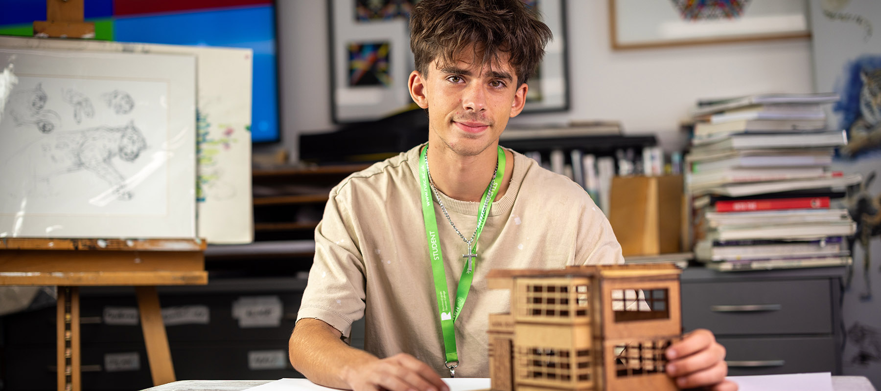 Male Richmond upon Thames College A-Level student showing his architecture model. He is sitting in the classroom with art books and drawings behind him. 