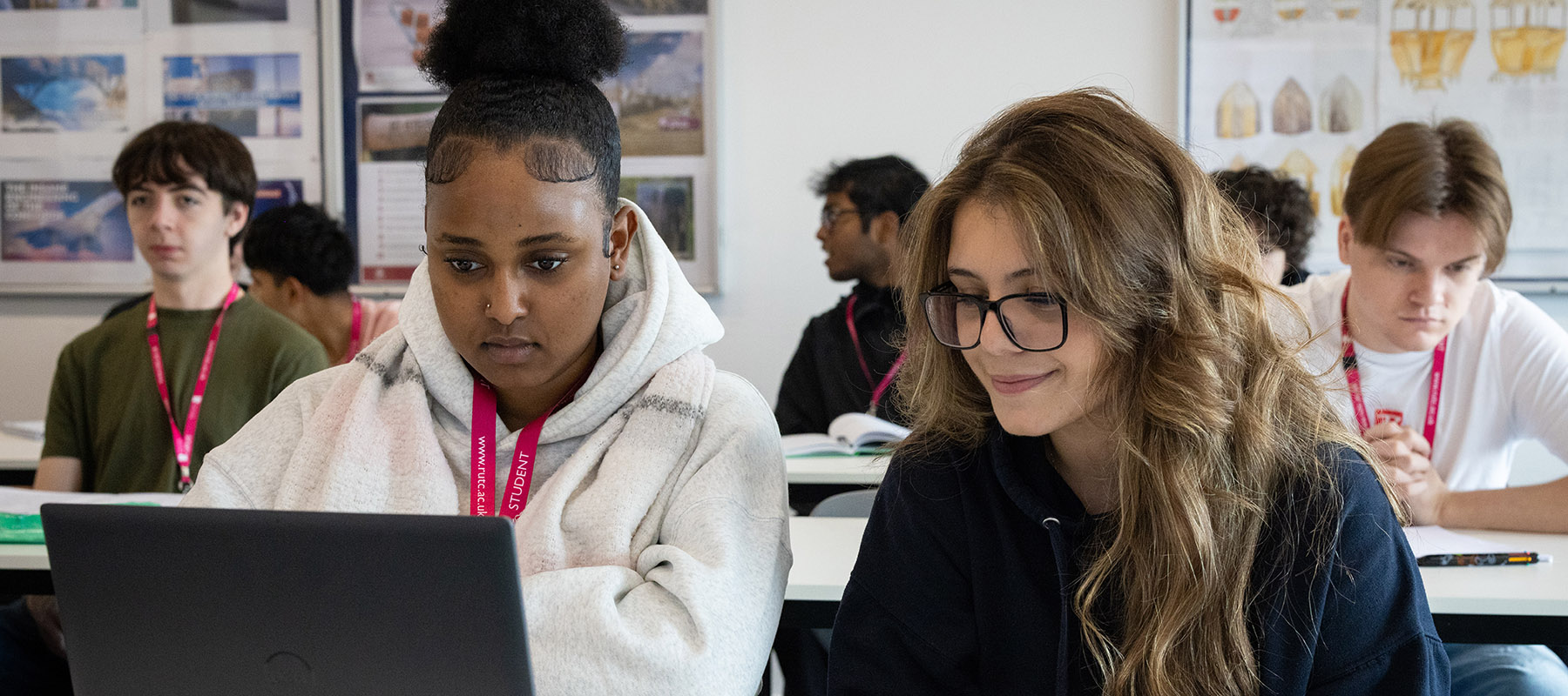 Two female Richmond upon Thames students looking at a laptop in class. One is wearing a white hoodie and one is wearing a navy hoodie and glasses. 