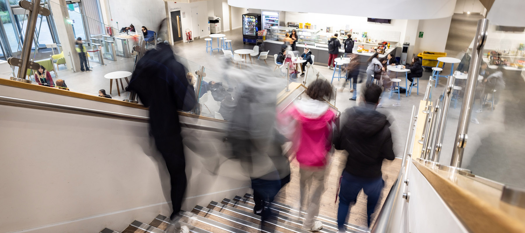 Blurred action shot of Richmond upon Thames College students walking down the stairs into the main reception, canteen area, which is buzzing with students. 