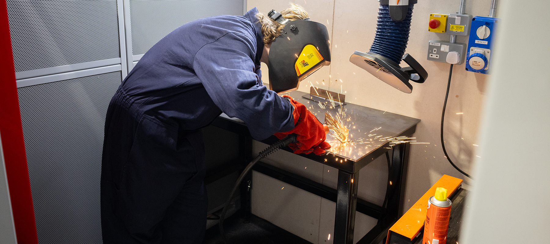 Richmond upon Thames student welding on a bench, wearing protective red gloves, helmet and blue overalls.