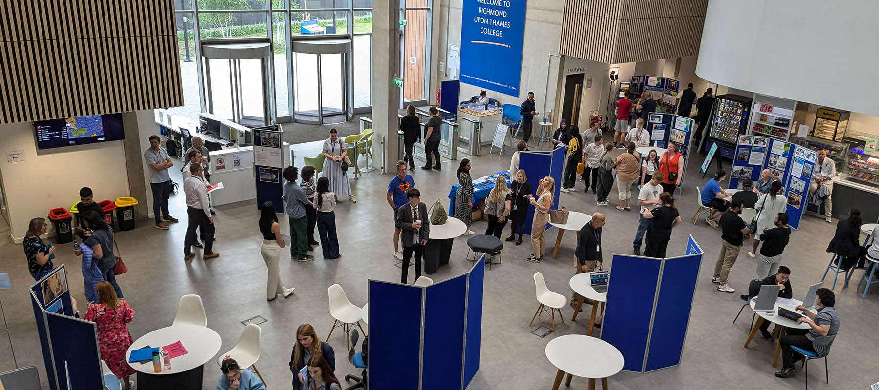 View from the balcony of one of the Richmond upon Thames College open events with people, booths and chairs scattered.