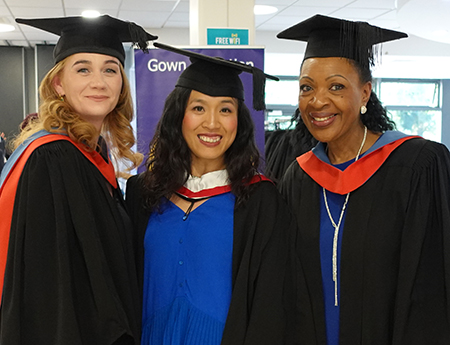 Three female Uxbridge staff members in their caps and gowns smiling at the camera and getting ready for the HE Graduation 2025. 