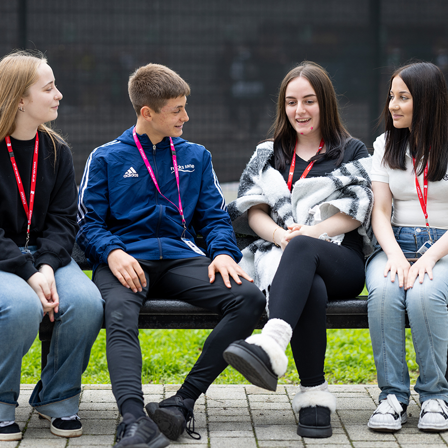A group of Richmond upon Thames students sitting on a bench and talking together outside the College. They are all wearing red or pink lanyards.