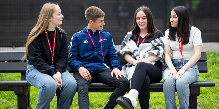 A group of Richmond upon Thames students sitting on a bench and talking together outside the College. They are all wearing red or pink lanyards.
