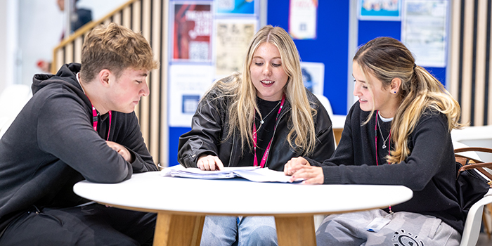 Three Richmond upon Thames students sitting around a table - talking and poinitng to a workbook. They are all dressed in black and wearing pink lanyards.
