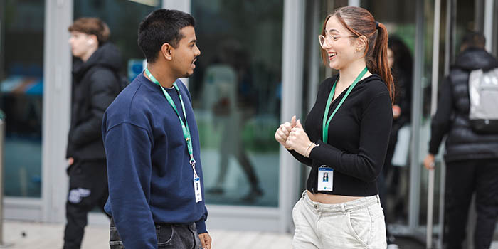 Richmond upon Thames College students talking to each other outside the main entrance to the College. There is one male and one female student, both wearing lanyards.