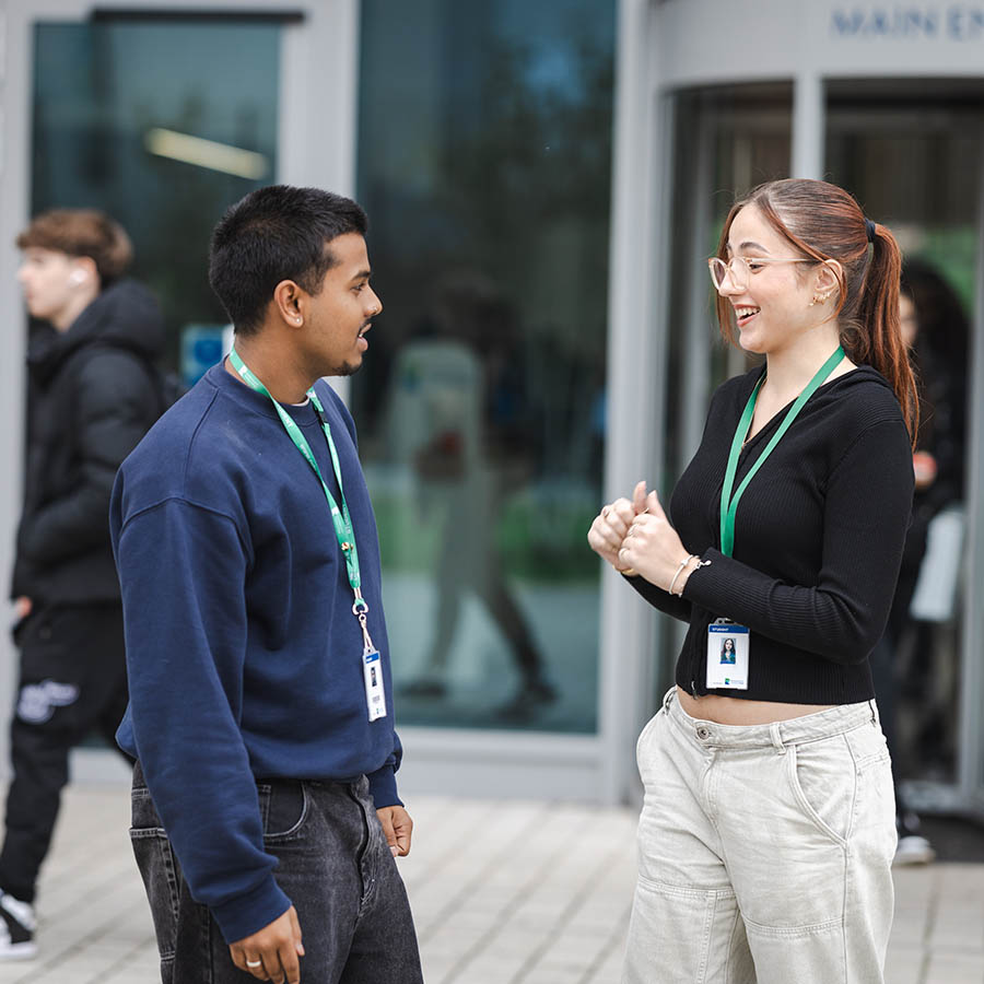 Richmond upon Thames College students talking to each other outside the main entrance to the College. There is one male and one female student, both wearing lanyards.