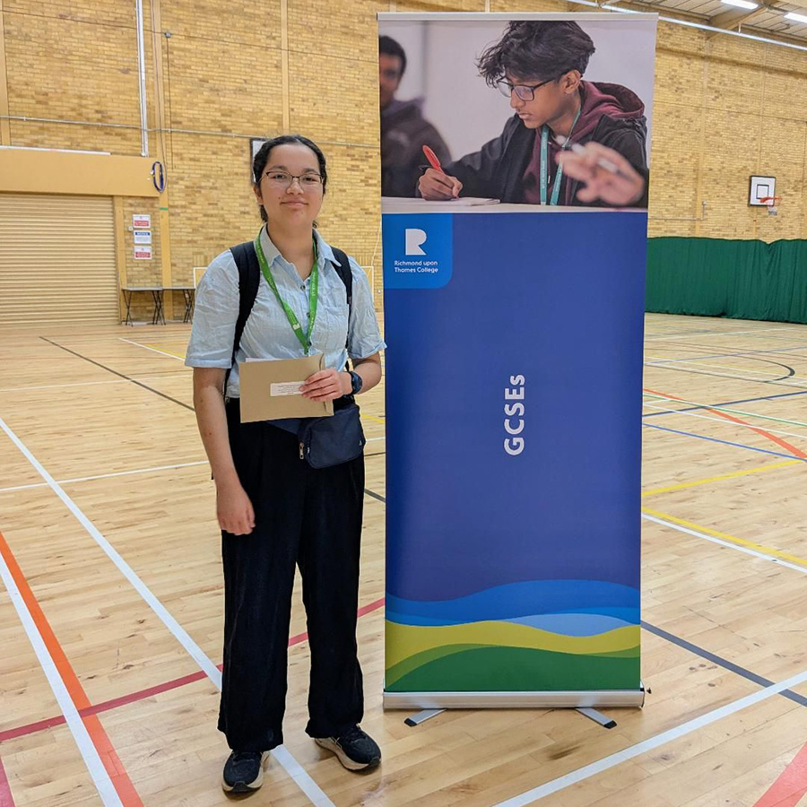 Miri - she is standing in the Richmond upon Thames Sports Hall next to a GCSE pull-up banner. She is wearing glasses, a green lanyard and smiling while holding her GCSE exam results. 