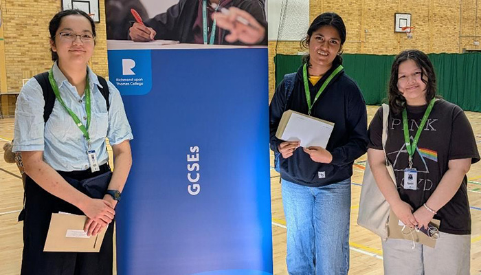 Miri and her two female friends standing in the Richmond upon Thames Sports Hall next to a GCSE pull-up banner. They are smiling and holding their GCSE exam reults. They are all wearing their green lanyards. 