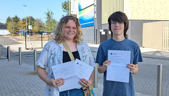 Two Richmond upon Thames College students holding their exam results and standing outside the College. The Sports centre is behind them and it is a sunny day.