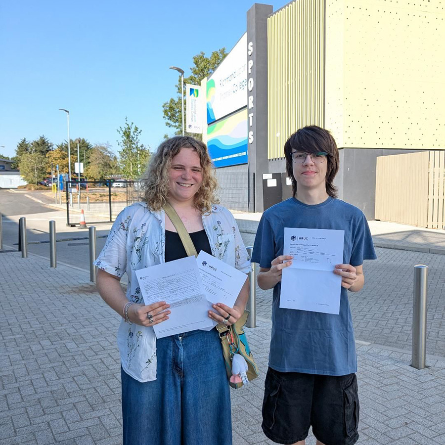 Two Richmond upon Thames College students holding their exam results and standing outside the College. The Sports centre is behind them and it is a sunny day.