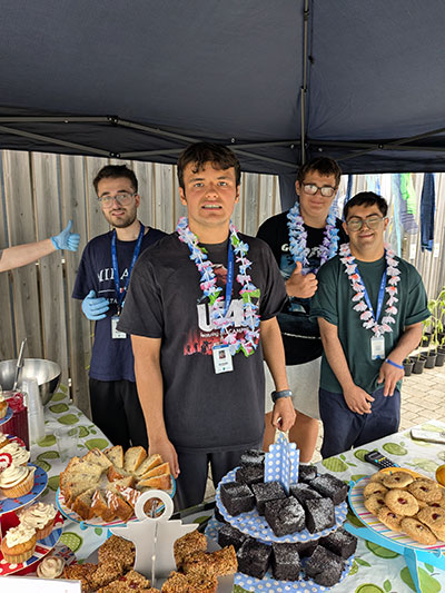 4 supported learning students selling cakes from a stand set up in the garden, all wearing flower garlands