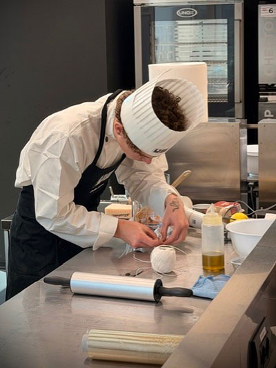 student, Charlie Sexton, in chef whites, in kitchen, leaning over work surface and creating something with kitchen utensils