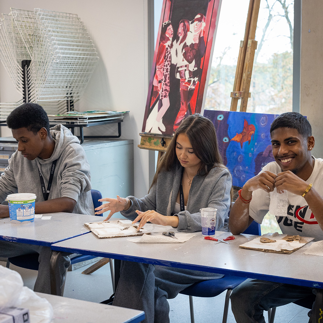 Richmond upon Thames College students sitting in an art studio working on their own art projects. One male student is smiling and looking at the camera.