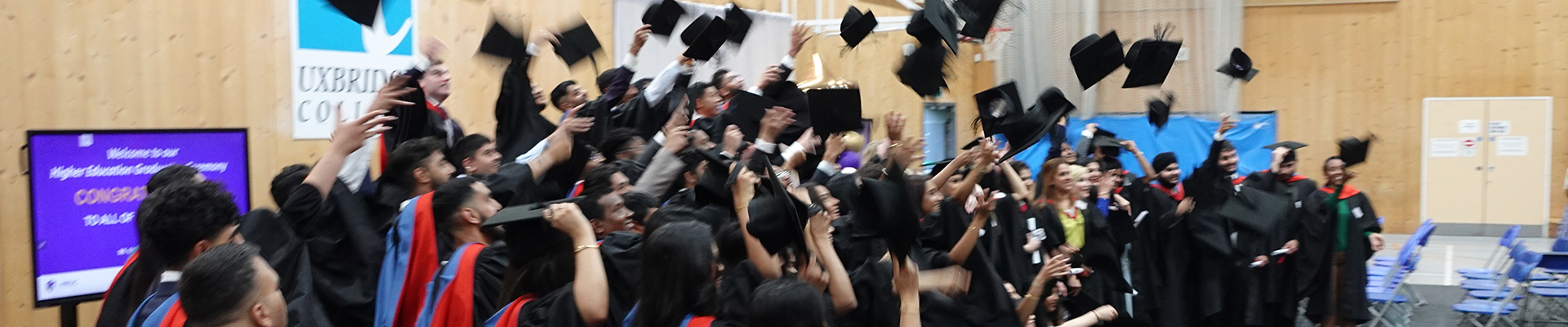 HE Students at their graduation in the Uxbridge College sports hall, throwing their black caps in the air. They are all wearing graduation robes.         
