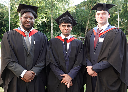 Three male HE graduation students in a group smiling at the camera, wearing their black caps and gowns with red and blue sashes. Trees are in the background.    