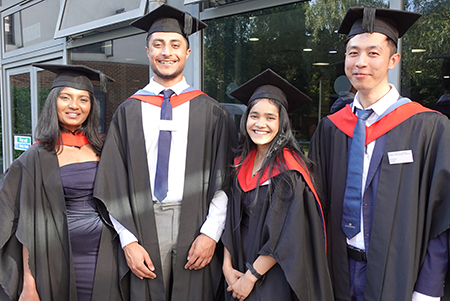 Two male and two female HE graduation students in a group smiling at the camera, wearing their black caps and gowns with red and blue sashes. Building windows are in the background.